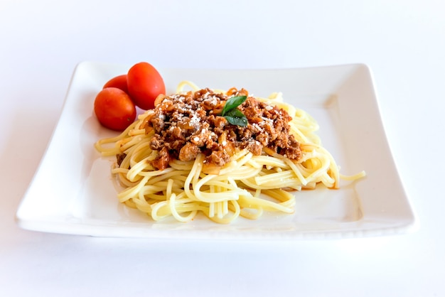 Spaghetti Bolognese podawane na białym talerzu na białym tle z pomidorami i bazylią.