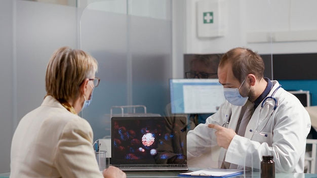 Specjalista ds. zdrowia pokazuje staruszce ilustrację wirusa na laptopie, wyjaśniając zagrożenie koronawirusem i diagnozę choroby. Emerytowany pacjent i lekarz z maską na twarzy podczas wizyty kontrolnej.