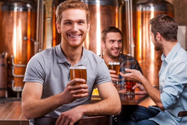Spędzanie czasu w pubie piwnym. Przystojny młody mężczyzna wznoszący tosty z piwem i uśmiechający się, podczas gdy jego przyjaciele rozmawiają w tle