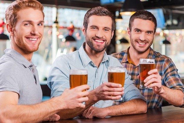 Spędzanie dobrego czasu w barze. Trzech szczęśliwych młodych mężczyzn w strojach codziennych, wznoszących tosty z piwem i uśmiechających się, siedząc razem przy barze?