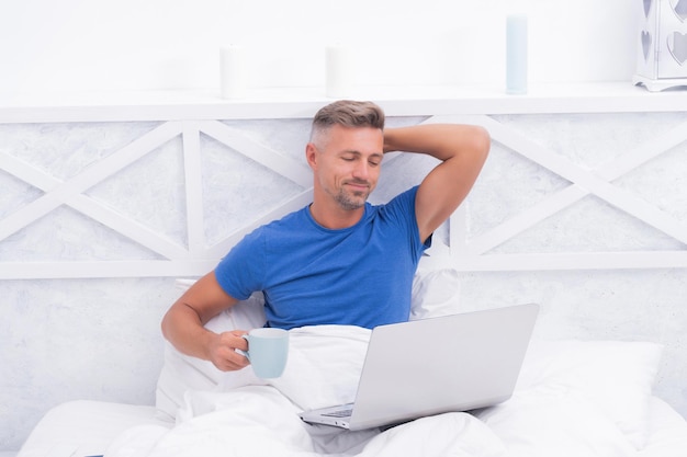 Spędzanie poranka w Internecie. Obudzony facet pić herbatę za pomocą laptopa. Rano w łóżku. Internetowe poszukiwanie relaksu. Czek na czacie.