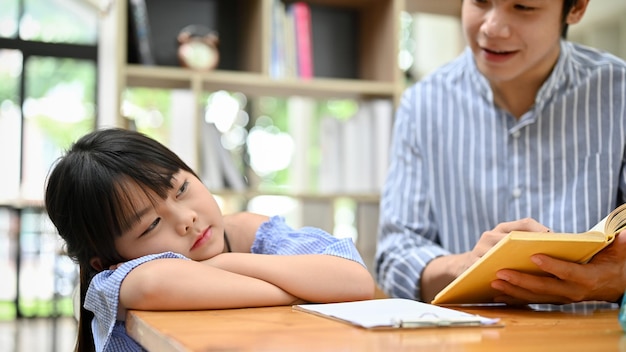 Śpiąca mała Azjatka opiera się o stół, podczas gdy jej nauczyciel uczy i zadaje pytanie