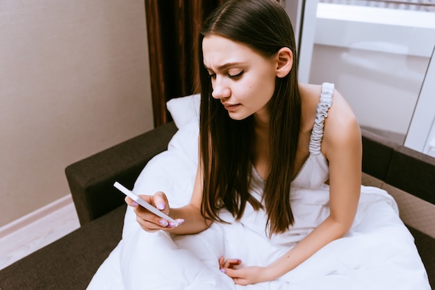 Śpiąca młoda dziewczyna siedzi w łóżku pod białym kocem, zagląda do swojego smartfona