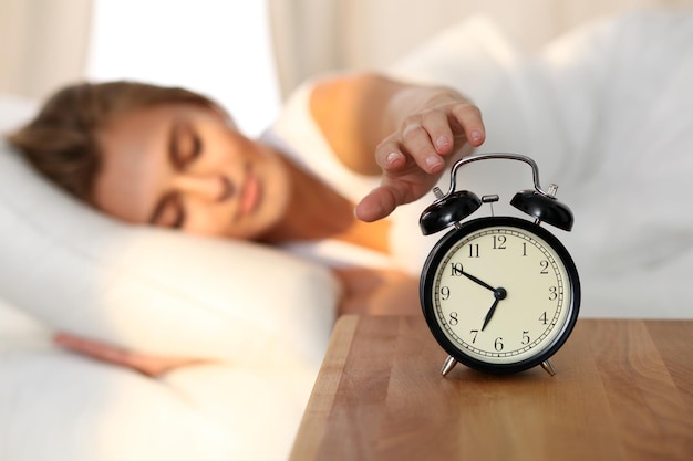 Śpiąca młoda kobieta wyciąga rękę do dzwoniącego alarmu, chcąc go wyłączyć Wczesne pobudki, niewystarczająca ilość snu, koncepcja pracy