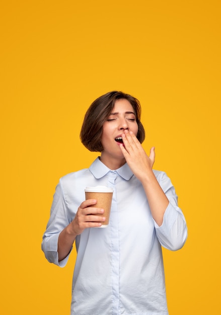 Śpiąca młoda kobieta z kubkiem gorącej kawy na wynos zakrywającym usta i ziewaniem z zamkniętymi oczami na żółtym tle rano
