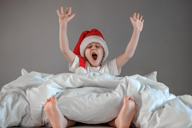Śpiący chłopiec siedzi na łóżku popijając w czapce Mikołaja Nowy Rok i Święta