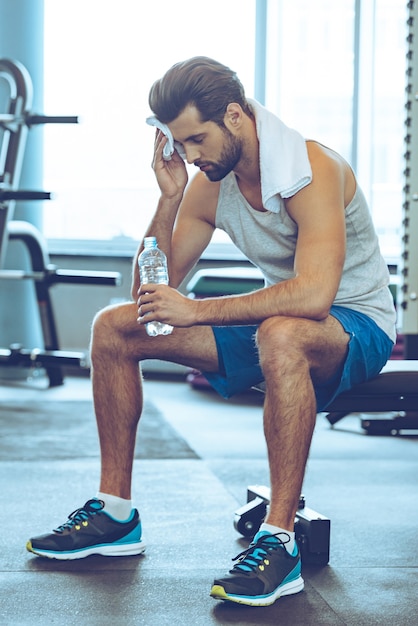 Spocony i zmęczony. Pełna długość przystojnych młodych mężczyzn w strojach sportowych, bijących pot ręcznikiem i trzymających butelkę wody podczas siedzenia na siłowni