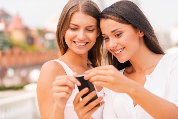 Spójrz na to! Dwie piękne młode kobiety patrzące na telefon komórkowy i uśmiechające się stojąc razem na zewnątrz