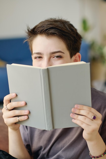 Spojrzenie uroczej nastolatki patrzącej w kamerę nad otwartą książką w szarej okładce