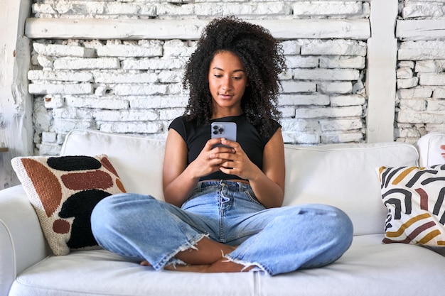 Spokojna Afrohaired kobieta używa telefonu na kanapie cieszy się miękkie światło poranne w domu