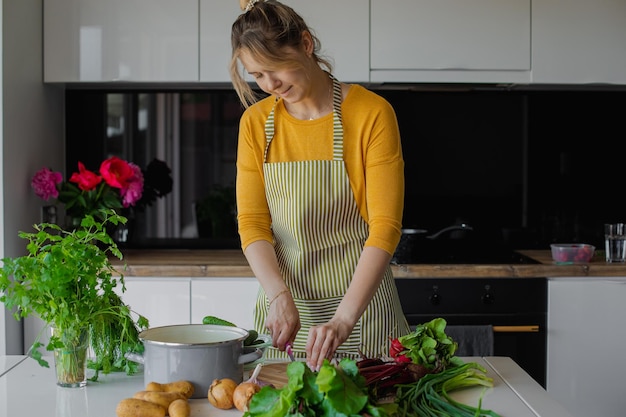 Spokojna i radosna blondynka w fartuchu sieka świeże warzywa i zieleń na pokładzie detox zdrowy