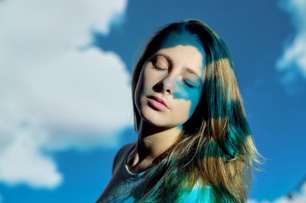 Spokojna młoda kobieta z długimi włosami zamykająca oczy pod projekcją jasnoniebieskiego nieba i białych chmur