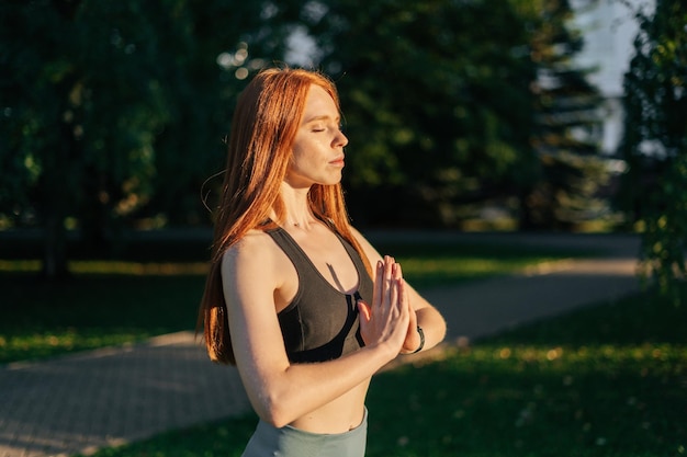 Spokojna młoda kobieta z zamkniętymi oczami wykonująca gest Namaste na zewnątrz w parku miejskim
