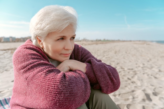 Spokojna, rozmarzona starsza pani siedząca na plaży i patrząca na coś w oddali?