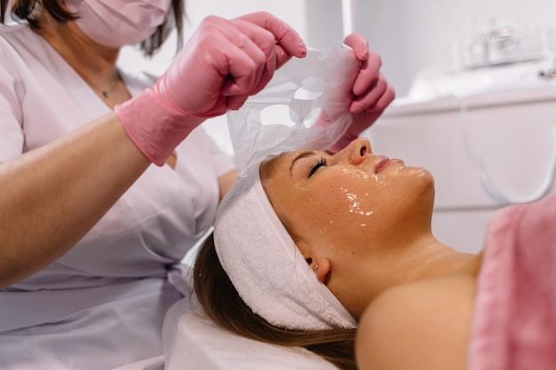 Spokojna spokojna kobieta leżąca podczas zabiegu kosmetycznego Ręce w białych rękawiczkach nakładają białą maskę na całą twarz do zabiegu kosmetycznego i pielęgnacji skóry