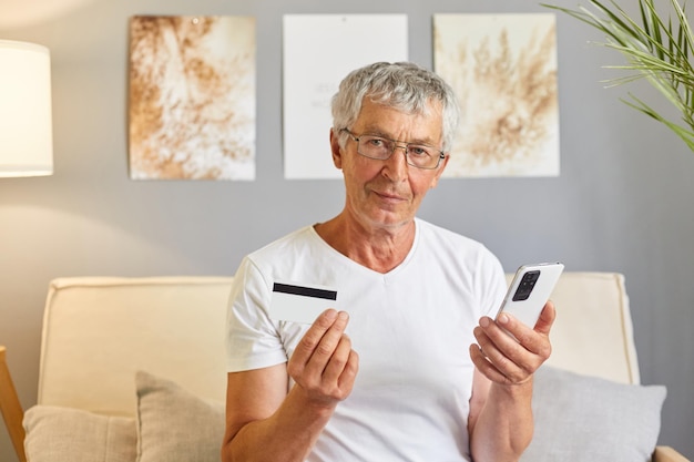 Spokojny dojrzały mężczyzna korzystający z telefonu komórkowego i dokonujący płatności online kartą kredytową klient robi zakupy z domu płacąc za zakupy w sklepach internetowych siedząc na kanapie w domu wnętrze