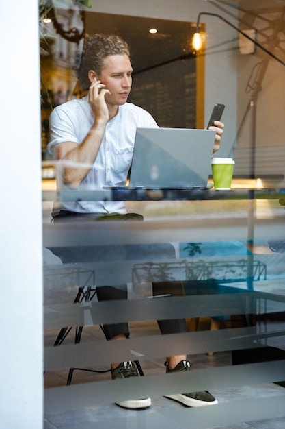 Spokojny kręcone biznes człowiek za pomocą smartfona siedząc przy stole z laptopem w kawiarni