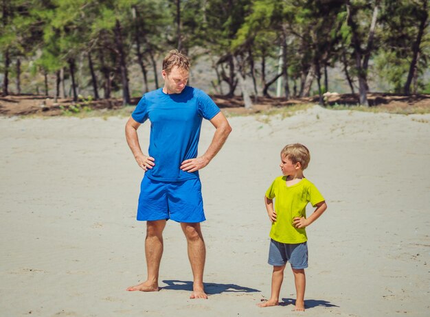 Sport dla małych dzieci Przystojny ojciec słodki syn ćwiczy na piaszczystej plaży w pobliżu leśnego parku po treningu ciesząc się razem na świeżym powietrzu prawidłowa technika kucania i zginania ręce na pasie