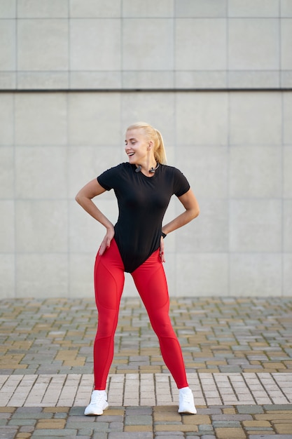 Sport i zdrowie. Atrakcyjna szczęśliwa szczupła dziewczyna ubrana w czarną koszulę, stylowe czerwone legginsy i białe trampki pozowanie na szarym tle ściany.