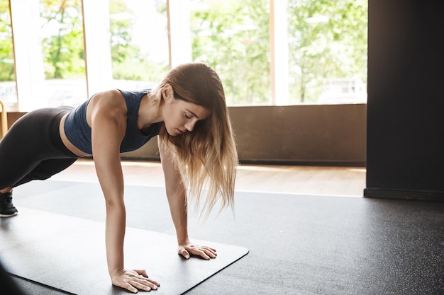 Sport Motywacyjny I Koncepcja Kobiet Skoncentrowana, Atrakcyjna Sportsmenka Z Długimi Włosami Nosi Sportowy Stanik I Legginsy Stojąc W Desce Gumowa Mata Pushup Podłoga Siłowni Podczas Aktywnej Sesji Treningowej