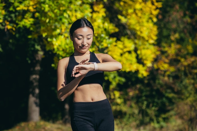 Sportowa azjatycka kobieta patrząca na inteligentny zegarek na nowoczesnym torze biegacza przy użyciu smartwatcha fitness bransoletka Portret fitness lekkoatletka sprawdzająca wynik jogging w odzieży sportowej w miejskim parku miejskim na zewnątrz