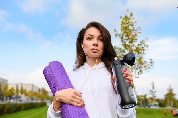 Sportowa brunetka kobieta na tle zielonego parku miejskiego z fioletową matą, nosić słuchawki, pić wodę po treningu, fitness, joga na zewnątrz