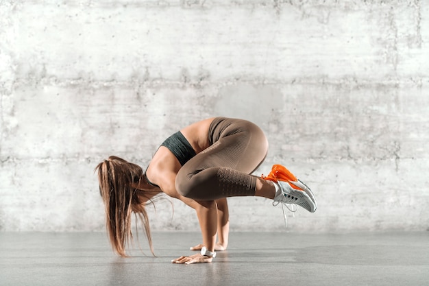 sportowa brunetka w kucyku i sportowym ubraniu, stojąca na rękach przed szarą rustykalną ścianą.