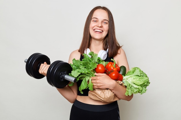Sportowa wysportowana kobieta podnosząca trzymając brzana i świeże warzywa izolowane na białym tle, ciesząc się zdrową żywnością ekologiczną i treningami