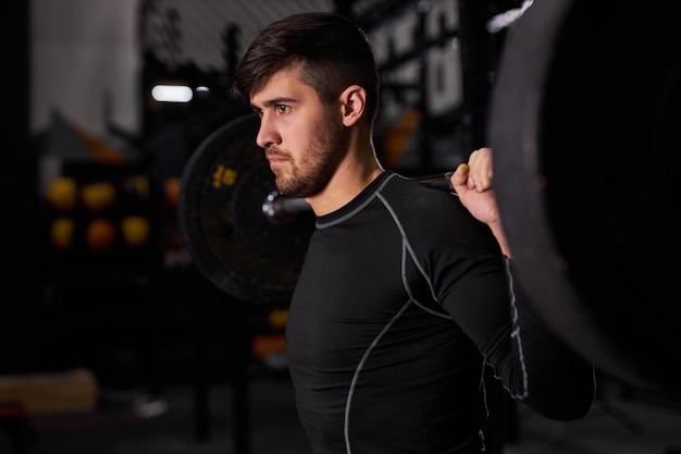 Sportowiec ćwiczący ze sztangą. Młody kaukaski muskularny mężczyzna kulturysta robi trening podnoszenia ciężarów w ciemnej siłowni, używając sprzętu sportowego, człowiek stoi patrząc z boku, widok z boku