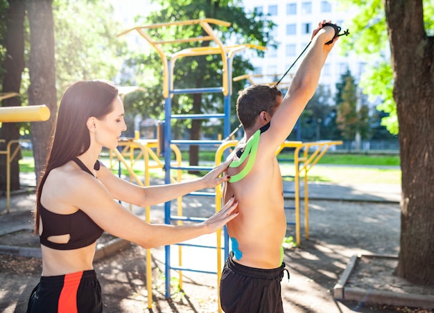 Sportowiec uśmiechnięta brunetka młoda kobieta instruktor pomaga przystojny mężczyzna w krótkich spodenkach z kinezjologią elastyczną taśmą na ramionach treningu ciała