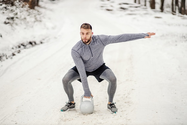 Zdjęcie sportowiec w kucki na zaśnieżonej ścieżce i podnoszący kettlebell fitness na świeżym powietrzu fitness zima kulturystyka