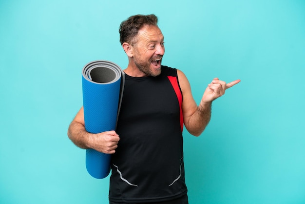 Sportowiec w średnim wieku idący na zajęcia jogi, trzymając matę odizolowaną na niebieskim tle, wskazując palcem na bok i prezentując produkt
