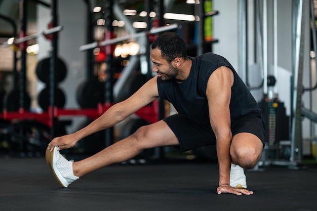 Sportowy młody czarny mężczyzna rozciągający mięśnie nóg podczas treningu na siłowni