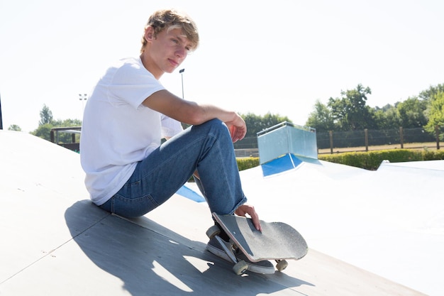 Sportowy nastolatek chłopiec siedzący na skate parku ze swoją deskorolką, robiący sobie przerwę i relaksujący się w słoneczny dzień