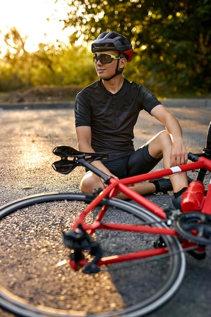 Sportowy rowerzysta w ubraniach rowerowych i kasku siedzi na drodze w pobliżu roweru, odpoczywając