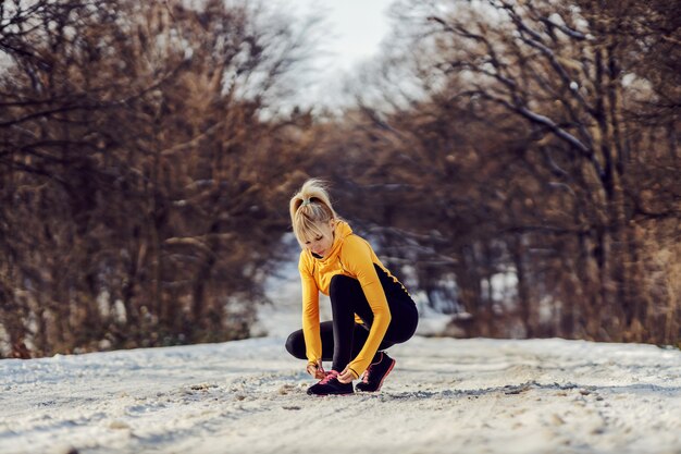 Sportsmenka przyczajona na zaśnieżonej ścieżce w przyrodzie w zimie i wiązanie sznurowadła. Odzież sportowa, fitness zimowy, zdrowe życie