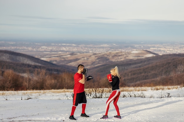 Sportsmenka sparing z rękawicami bokserskimi w przyrodzie w śnieżny zimowy dzień ze swoim trenerem. Boks, fitness zimowy, fitness na świeżym powietrzu