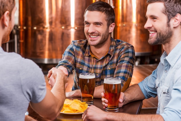 Spotkanie ze starymi przyjaciółmi. Trzech szczęśliwych młodych mężczyzn siedzi razem w pubie piwnym, podczas gdy dwóch z nich ściska dłonie