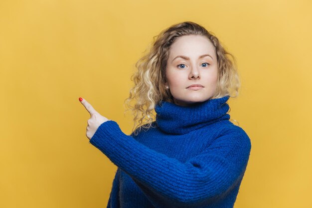 Sprawdź to Poważna blondynka, młoda kobieta z kręconymi blond włosami, ubrana w jasnoniebieski sweter, wskazuje na puste miejsce na tekst promocyjny lub reklamę Pusta żółta ściana
