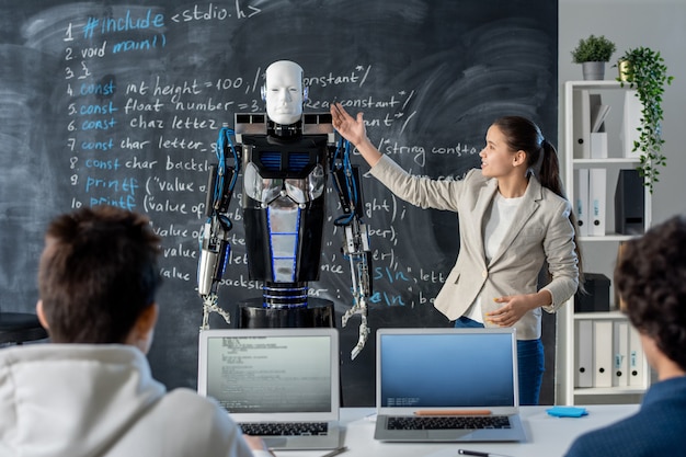 Sprytna dziewczyna stojąca przy tablicy i wskazująca na robota, przedstawiając kolegom z klasy jego cechy charakterystyczne na seminarium