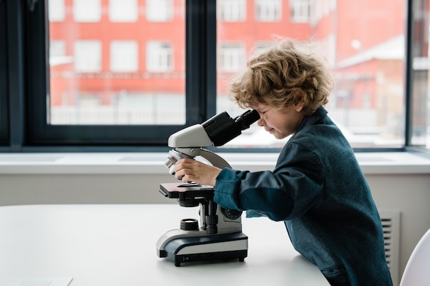 Sprytny uczeń lub biolog w codziennym ubraniu pochyla się nad biurkiem, patrząc pod mikroskopem na okno laboratorium