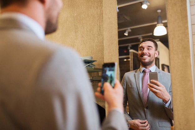 sprzedaż, zakupy, moda, styl i koncepcja ludzi - szczęśliwy młody człowiek lub biznesmen w garniturze ze smartfonem robiący lustrzane selfie w sklepie odzieżowym