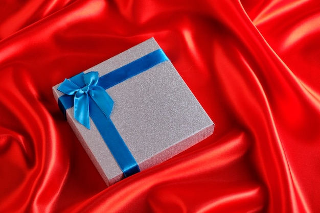 Srebrne pudełko z niebieską wstążką i kokardką na jedwabnym tle z czerwonej tkaniny