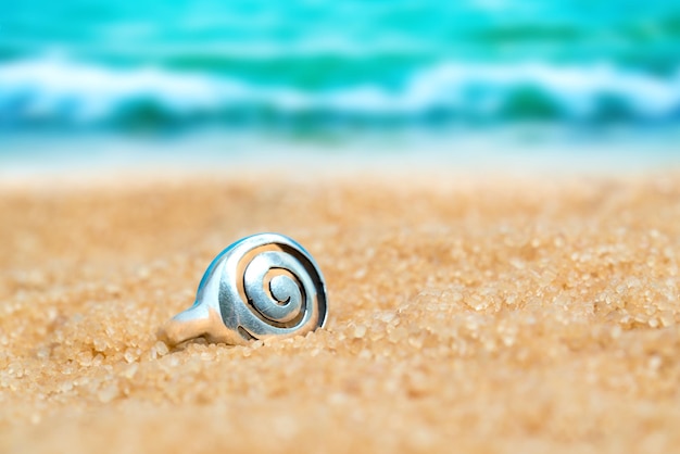 Srebrny pierścionek ze spiralnym wzorem w piasku na tle plaży i morza