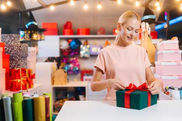Zdjęcie Średnie ujęcie portret młodej kobiety sprzedającej pakującej świąteczne pudełko na prezent wiązanej czerwoną wstążką i dekor...