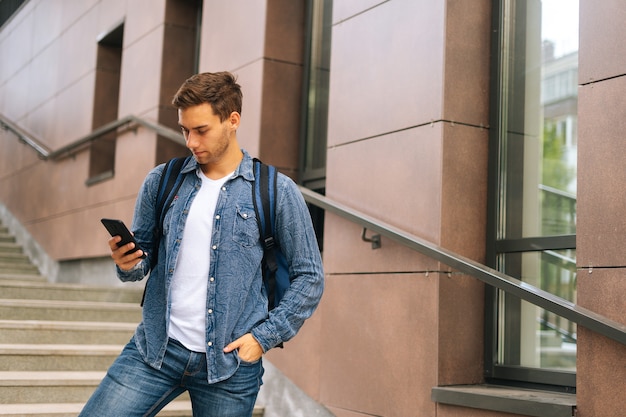 Średnie ujęcie przystojnego mężczyzny dostawy z dużym termo plecakiem trzymającym telefon, patrząc na ekran, za pomocą aplikacji nawigacyjnej. Kurier mężczyzna coraz zamówienia stojący na schodach budynku biurowego.