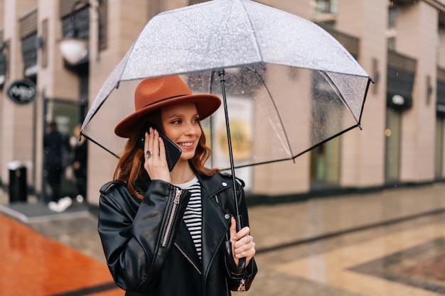 Średnie ujęcie szczęśliwej młodej kobiety w modnym kapeluszu stojącej z przezroczystym parasolem rozmawiającej na smartfonie w deszczu na europejskiej ulicy miasta