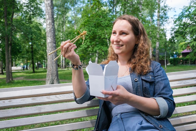 Średnio dorosła dojrzała kobieta na spacerze w letni dzień w parku je lunch z wokiem z makaronem