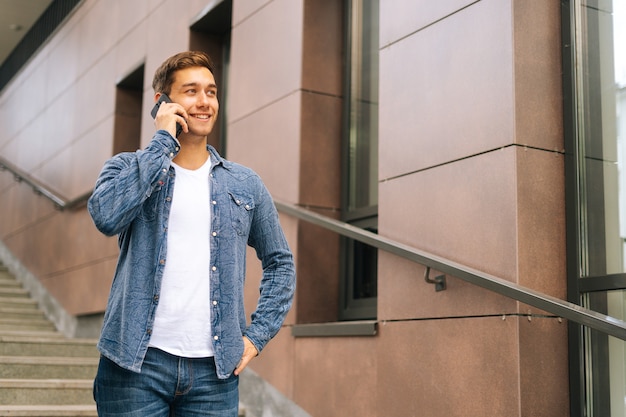 Średnio strzał portret pewnie przystojny młody mężczyzna rozmawia przez telefon komórkowy stojący na schodach nowoczesnego biurowca. Sukcesy biznesmen mówiąc na smartfonie na zewnątrz.