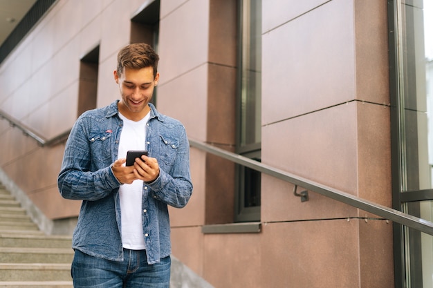 Średnio strzał portret wesoły przystojny młody człowiek za pomocą telefonu komórkowego stojącego na schodach nowoczesnego biurowca. Radosny mężczyzna ubrany w stylową odzież dżinsową czytanie wiadomości na zewnątrz.